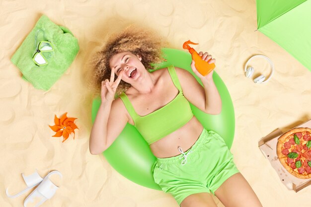 woman with curly hair lies on green inflatable swim ring holds bottle of suncream makes peace gesture has fun at beach eats pizza different items around enjoys good summer rest