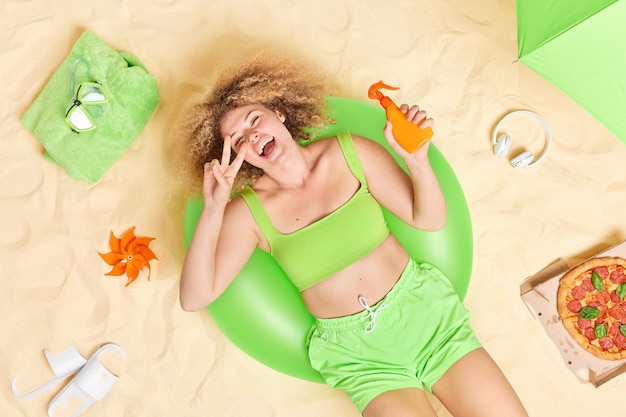 곱슬머리를 한 여성이 녹색 팽창식 수영 링에 누워 선크림 한 병을 들고 평화 제스처를 취하게 하고 해변에서 즐겁게 지냅니다. 피자를 먹습니다.