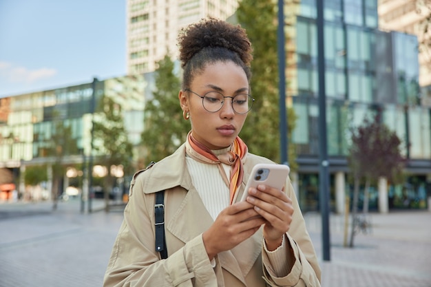 巻き毛の女性が現代のスマートフォンデバイスのチャットを注意深く見ていますインターネットで買い物をするのは丸い眼鏡をかけ、レインコートはダウンタウンを散歩します