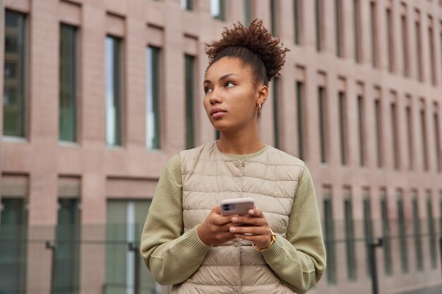 巻き毛の女性が目をそらし、余暇の間に外を思慮深く散歩する無料のインターネット接続を使用して、都市の建物に対してジャンパーとベストのポーズを着たアプリをインストールします