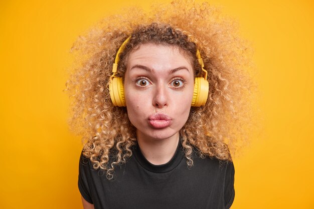 женщина с вьющимися волосами смотрит в камеру с кокетливым выражением лица, держит губы сложенными, слушает музыку в наушниках, наслаждается свободным временем, одетая в повседневные позы футболок