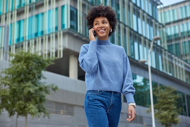 woman with curly hair makes call via smartphone expresses positive emotions wears casual blue jumper and jeans poses against modern glass building feels good during spare time