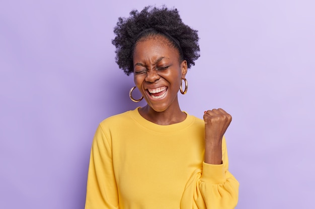  woman with curly hair makes yes gesture feels like winner raises fist wears yellow jumper and earrings isolated on purple 