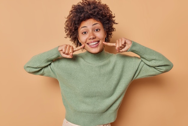 Free photo woman with curly hair points at smile shows white perfect teeth dressed in casual jumper isolated on beige being in good mood. happy human face expressions