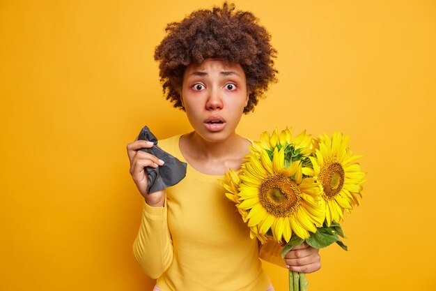 巻き毛の赤い腫れた目を持つ女性は、鮮やかな黄色で屋内でポーズをとっているヒマワリに対するアレルギーのためにナプキンのくしゃみを保持します