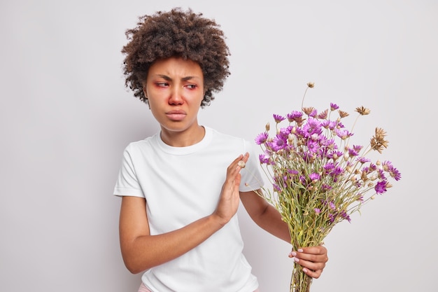 花粉にアレルギーのある野生の花の花束を手に入れることを拒否する巻き毛の女性は不幸に見えます赤い目と鼻は白い壁にさりげなくポーズをとっています