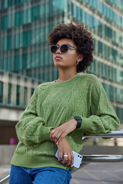 woman with curly hair rests after long walk in city wears sunglasses green jumper and jeans holds mobile phone waits for call