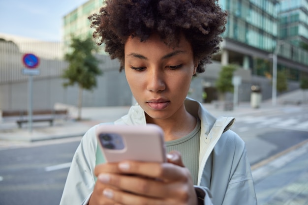 woman with curly hair sends message on smartphone uses cellphone app strolls against urban street dressed in casual clothes focused in screen of cellular