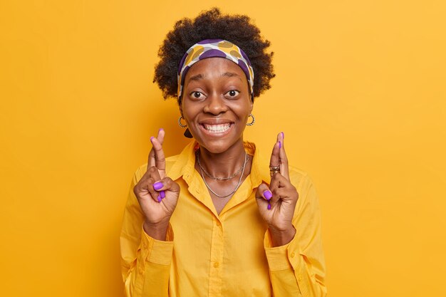  woman with curly hair smiles broadly keeps fingers crossed believes in good luck wears headband and casual shirt isolated on vivid yellow 