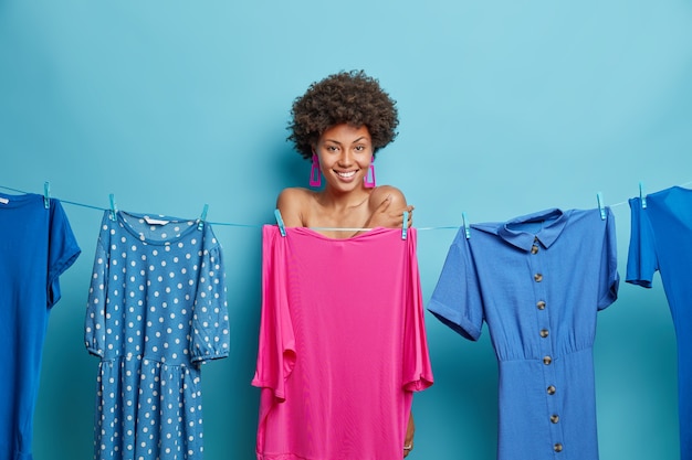 woman with curly hair stands shy hides naked body behind dress on clothesline smiles gladfully going to get dressed on special event isolated on blue