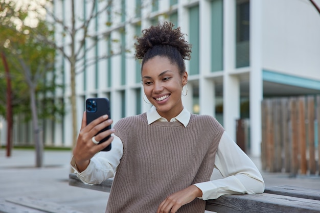 woman with curly hair talks via video call holds mobile phone dressed in stylish clothes makes photo of herself poses at city expresses positive emotions greets friend distantly