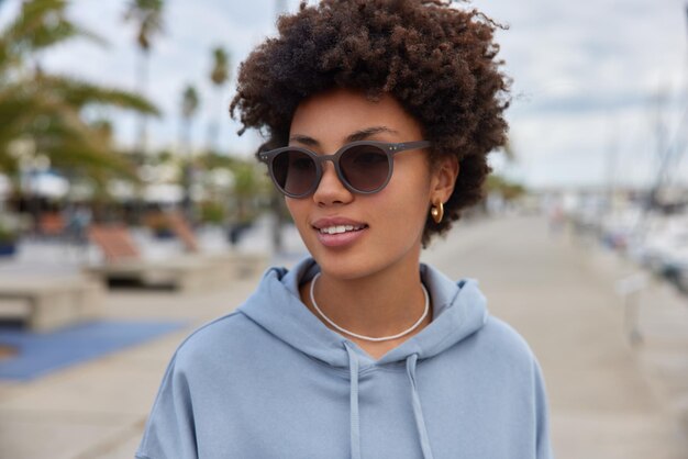 woman with curly hair wears sunglasses and hoodie walks at pier harbor admires scenic views. Female tourist on shore strolls during daytime. Tourism and recreation concept
