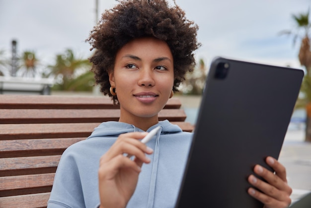 woman with cutly hair holds modern tablet and stylus creats pictures or edits photos works outdoors wears casual hoodie poses at wooden bench duing daytime focused somewhere