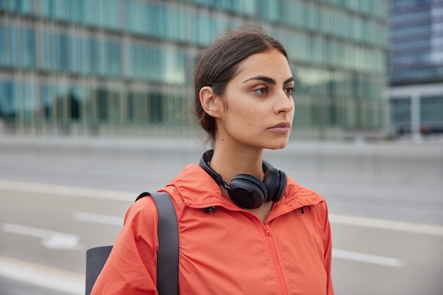  woman with dark combed hair looks thoughtfully into distance wears windbreaker carried karemat for training uses stereo headphones to listen music during workout
