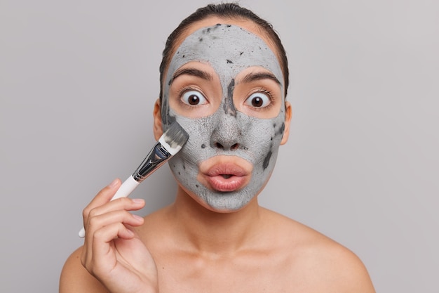 женщина с темными волосами наносит глиняную пилинг-маску на лицо, держит косметическую кисть, смотрит с большим удивлением при посещении камеры спа-салон стоит топлесс на серой стене