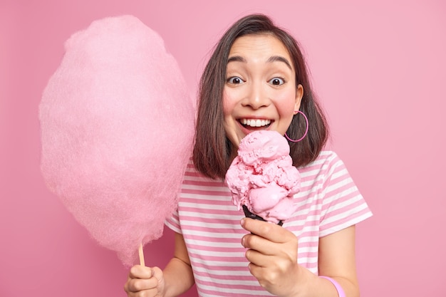 黒髪の女性がコーンの食欲をそそるアイスクリームを噛むスティックに甘い綿菓子を保持カジュアルなストライプのtシャツを着て楽しんでいます