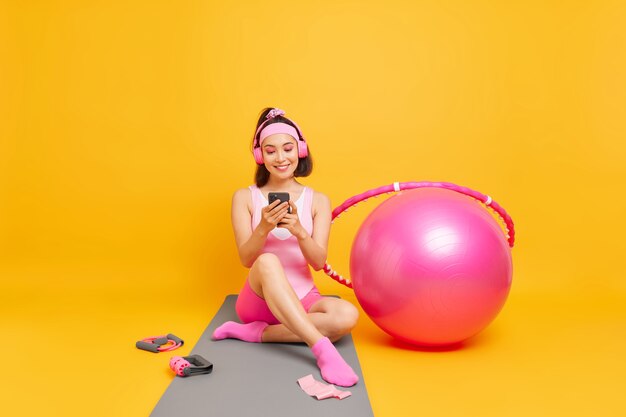 woman with dark hair checks her sport achievements on smartphone application sits on fitness mat dressed in activewear uses swiss ball hula hoop goes in for sport poses indoor