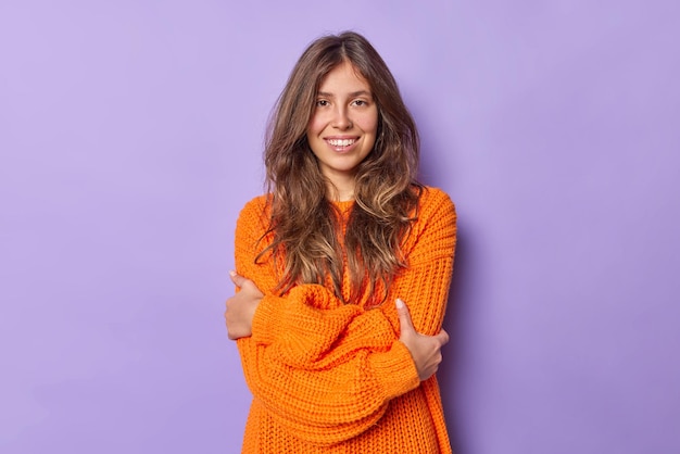 woman with dark hair embraces herself wears warm knitted orange sweater smiles positively poses on purple feels chilly walking during cold weather