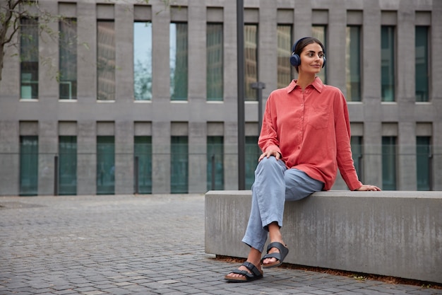 Donna con i capelli scuri indossa abiti casual comodi distoglie lo sguardo ascolta musica in cuffie pose su lastra di pietra vicino a un edificio moderno. stile di vita delle persone e concetto di hobby