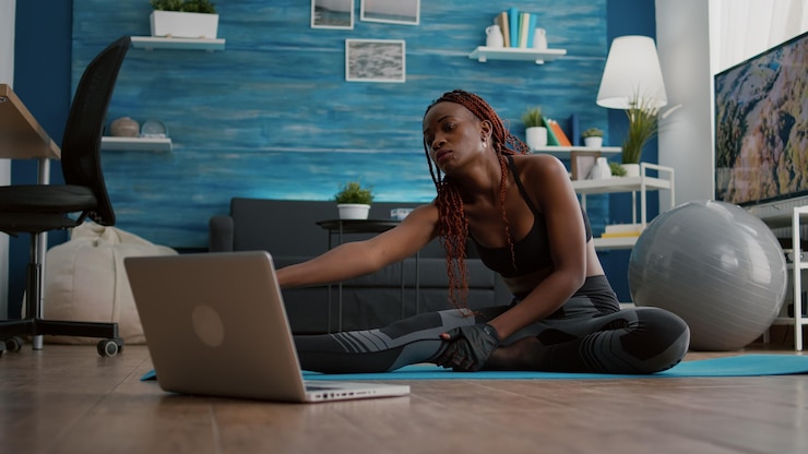 Free Photo woman with dark skin doing pilates workout in living room stretching body muscles on yoga map while watching online fitness sport video on laptop. flexible adult enjoying healthy lifestyle
