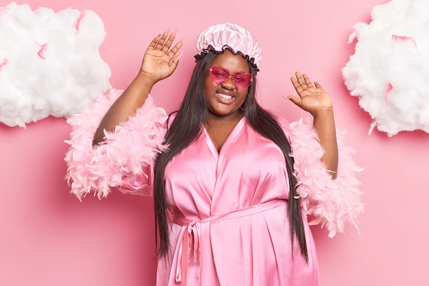 woman with dark skin smiles pleasantly raises arms dances carefree wears bath hat dressing gown and sunglasses isolated on pink