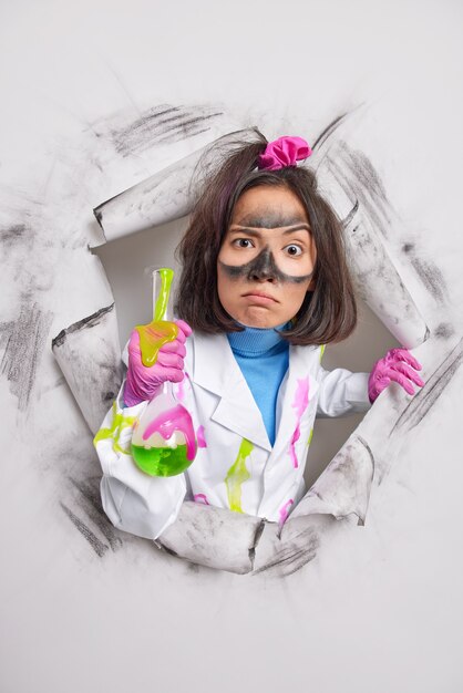  woman with dirty face holds flask with colorful liquid wears white coat rubber gloves breaks through paper 