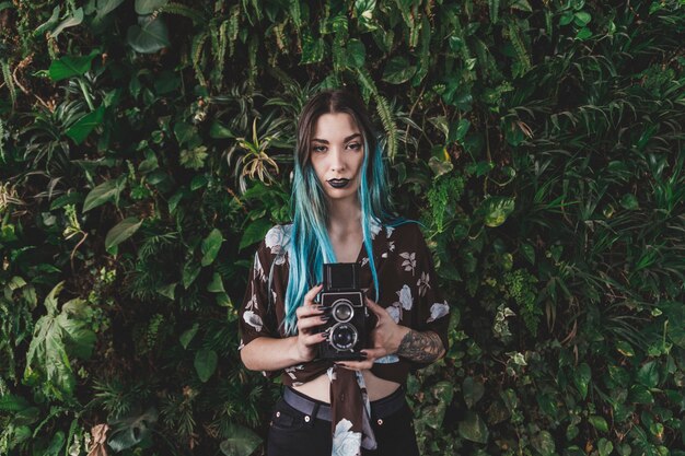 Woman with dyed hair holding retro styled camera in front of plant