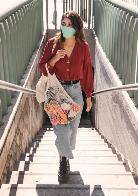 Woman with face mask climbing stairs while holding grocery bags