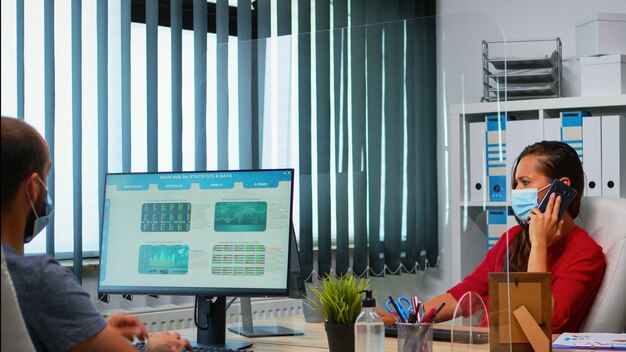 Woman with face mask talking on phone looking at desktop and analyzing the statistics. Freelancer working in workplace chatting with remotely team speaking on smartphone in front of computer