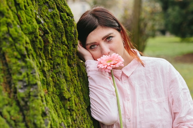 Женщина с цветком возле лица, опираясь на дерево в парке