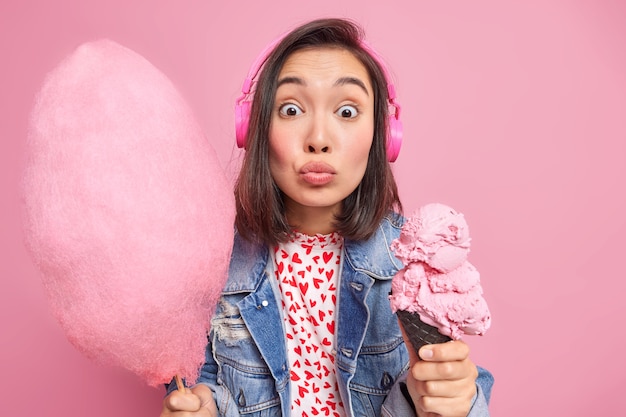 woman with folded lips listens music via wireless headphones holds tasty cotton candy and ice cream in waffle dressed in fashionable clothes