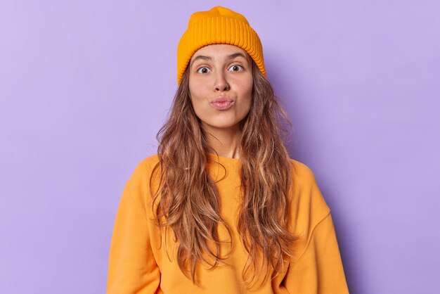 woman with folded lips looks surprisingly at camera has flirty expression wears casual jumper and hat isolated on purple blows air kiss makes smack gesture