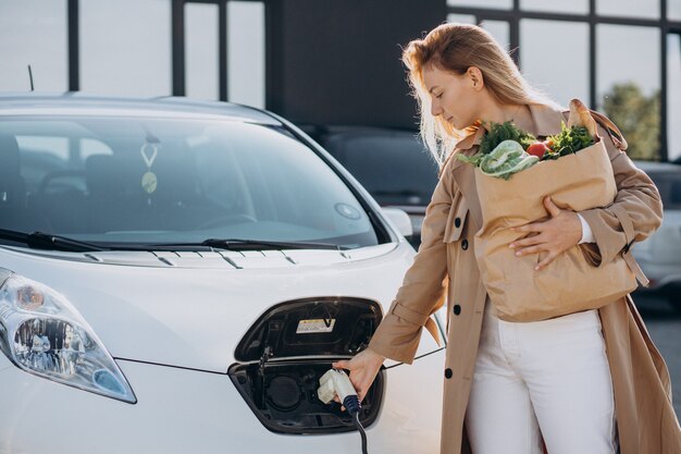 Женщина с сумками для покупок с едой заряжает электромобиль