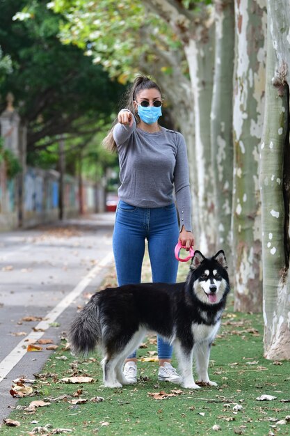Женщина в очках и хирургической маске держит собаку на поводке, указывая на камеру