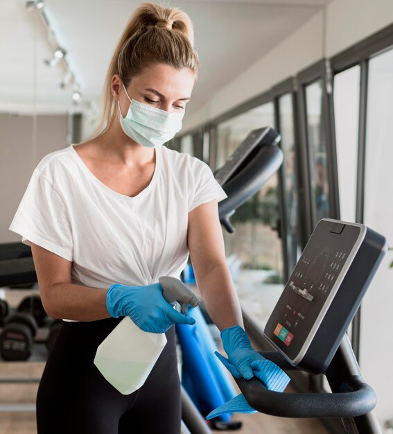 woman with gloves cleaning gym equipment while wearing medical mask