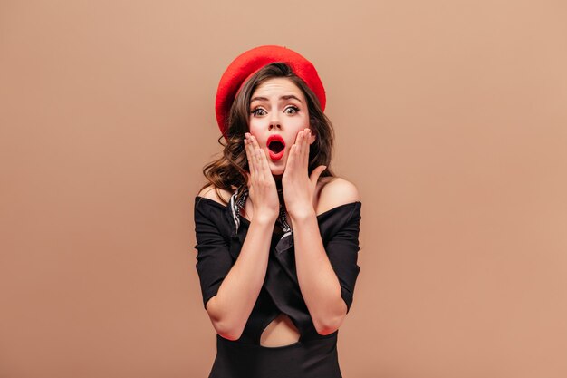 Free Photo  Girl in black dress and red beret stares into camera in  amazement and covers her mouth with hand.