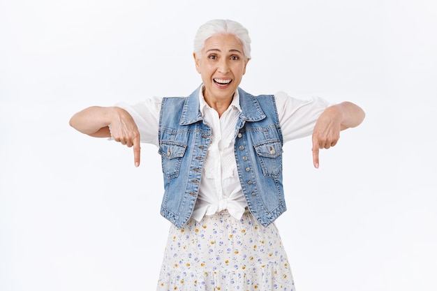 Free photo woman with grey combed hair, wear denim vest, dress, pointing fingers down, smiling and laughing amused, look camera interested and excited