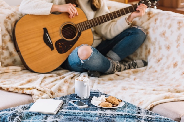 スマートフォン、メモ帳、ドリンクとクッキーのカップでテーブルの近くにギターを持つ女性