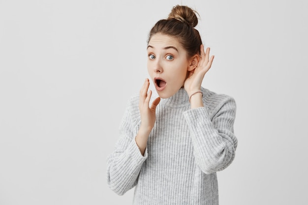 Woman with hair in bun listening to something holding hand near her ear. Curious girl being attentive trying to overhear some news. Curiosity concept