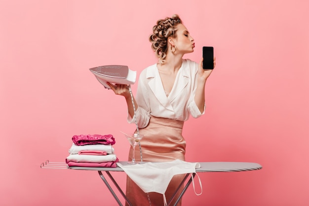 Woman with hair curlers holds iron and kisses her smartphone