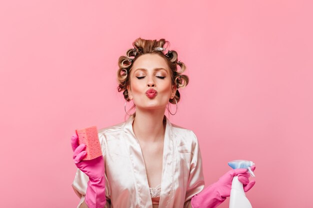 Бесплатное фото Женщина с бигуди на голове держит губку для мытья посуды и посылает воздушный поцелуй на розовую стену