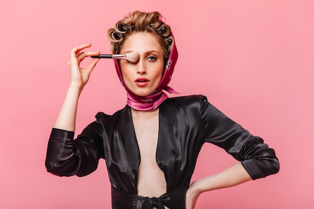 woman with hair curlers poses on pink wall and covers eye with makeup brush