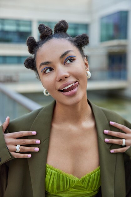 woman with hairstyle sticks out tongue dressed in fashionable clothes thinks about something pleasant applies blue eyeliner poses has good mood