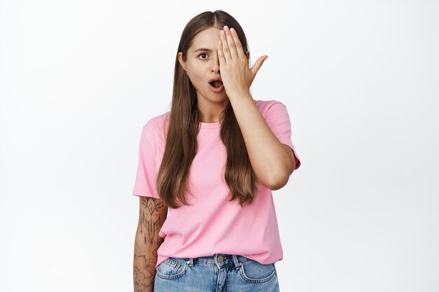 Woman with hand on half of face, looks with one eye with shocked, worried expression, standing against white background.