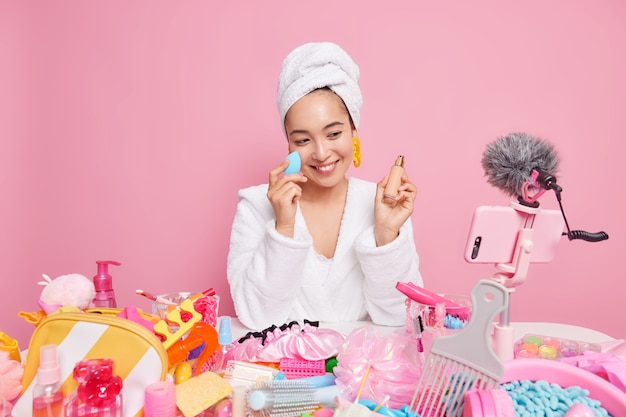 woman with happy expression applies foundation on face uses socail media for marketing records video for her beauty blog poses at smartphone camera isolated on pink wall. Online translation