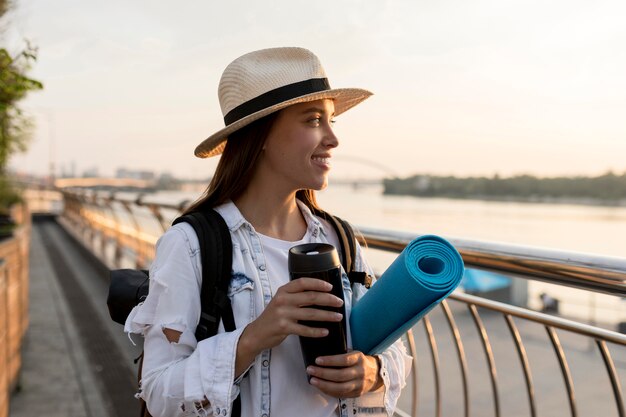 Женщина в шляпе и рюкзаке держит термос во время путешествия