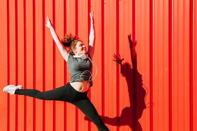 Foto gratuita donna con le cuffie che salta vicino al muro rosso