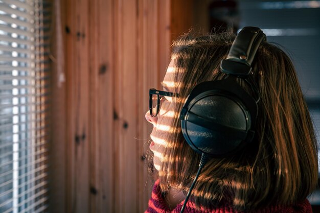 A woman with headphones looks through the blinds at the early morning sunlight