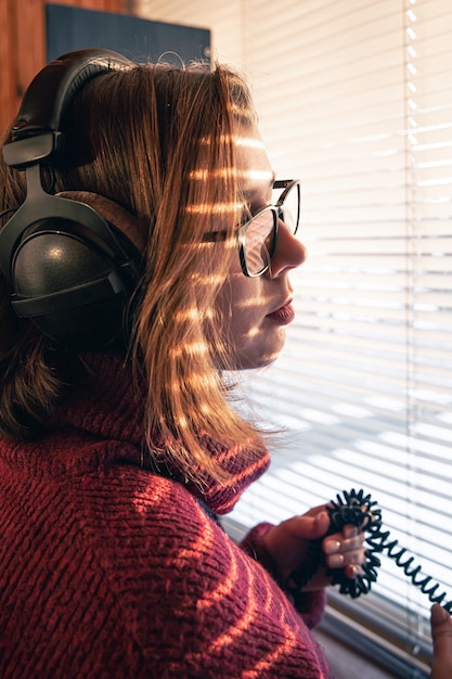 A woman with headphones looks through the blinds at the early morning sunlight