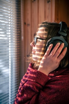 A woman with headphones looks through the blinds at the early morning sunlight